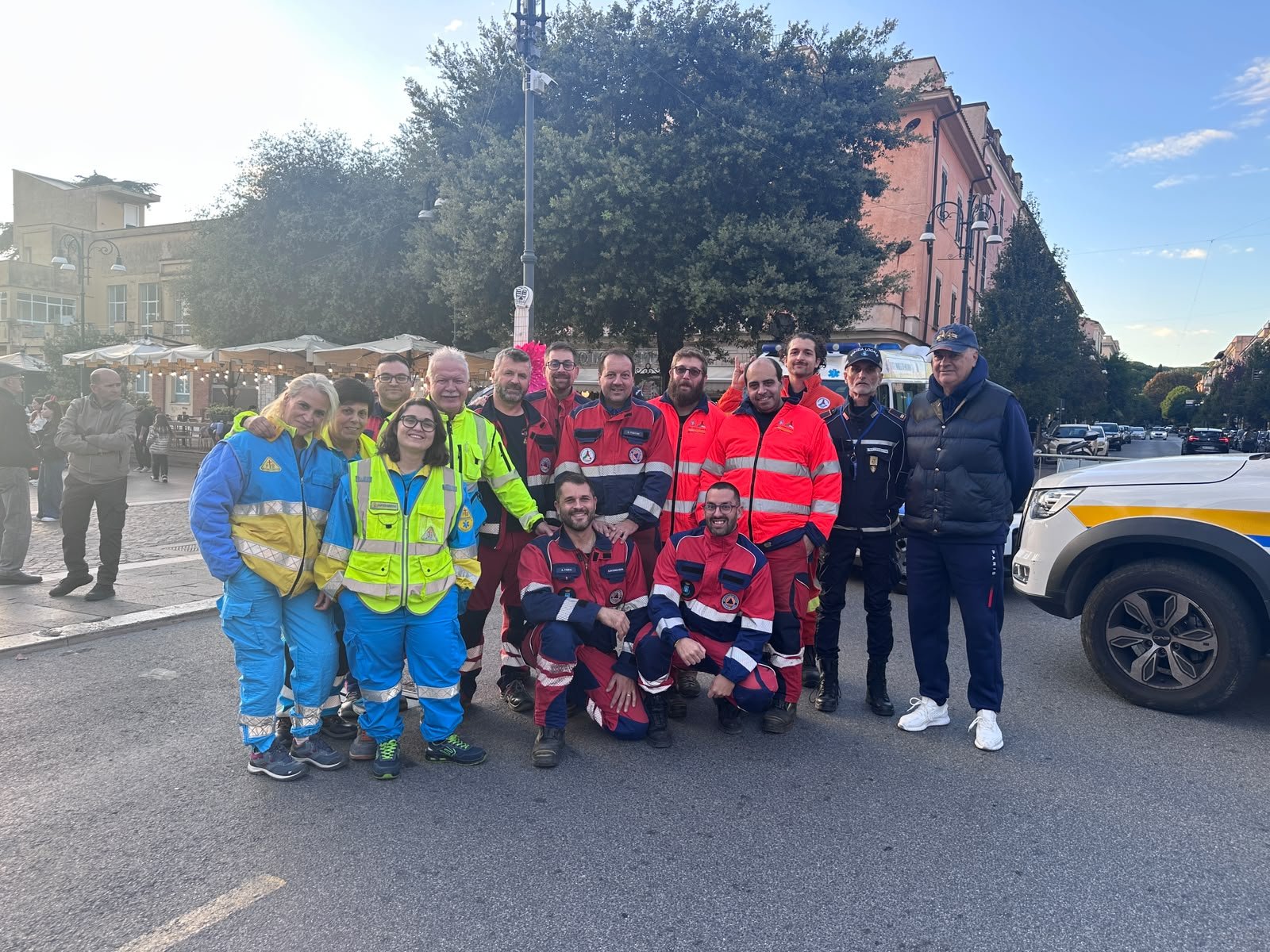 La squadra in posa in piazza: volontarie e volontari della Misericordia di Ariccia nelle divise blu e gialle, volontari della Protezione Civile di Genzano di Roma e un agente della Polizia Locale.