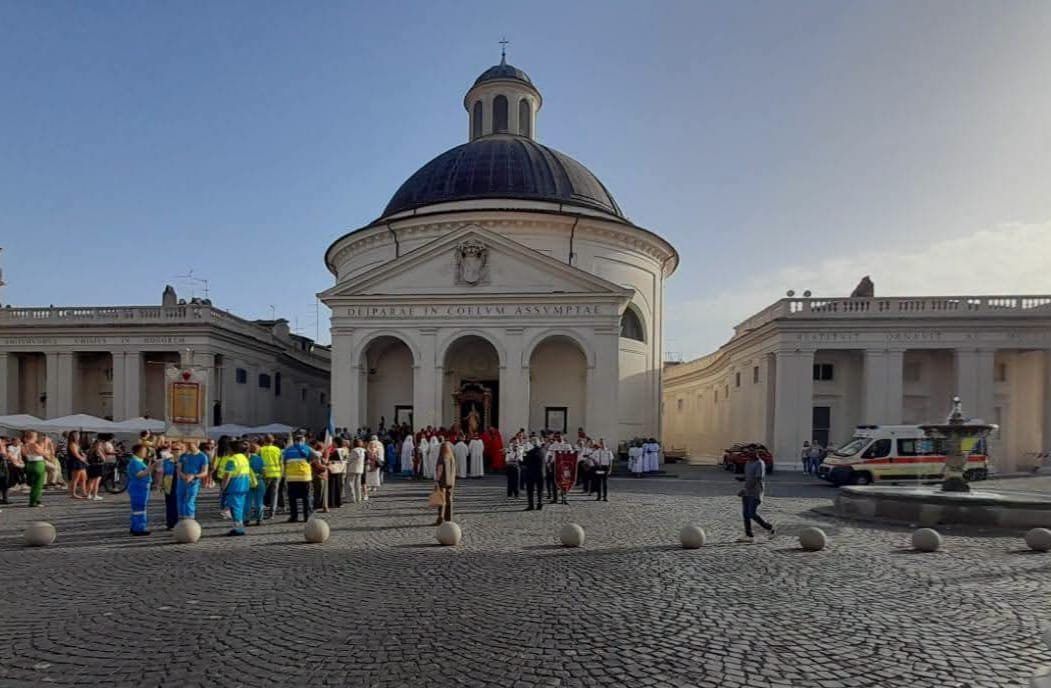 Piazza di Corte con la chiesa di Santa Maria Assunta, la processione in formazione e l'ambulanza della Misericordia accanto alla fontana