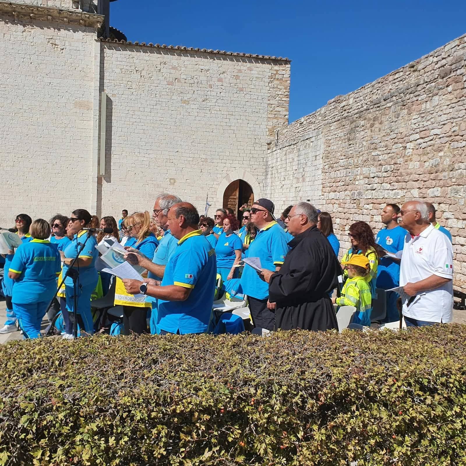 I volontari delle Misericordie di tutta Italia, in divisa azzurra e gialla, cantano e pregano con i libretti in mano davanti alle mura medievali di Assisi, sotto il sole; tra loro un frate in saio nero