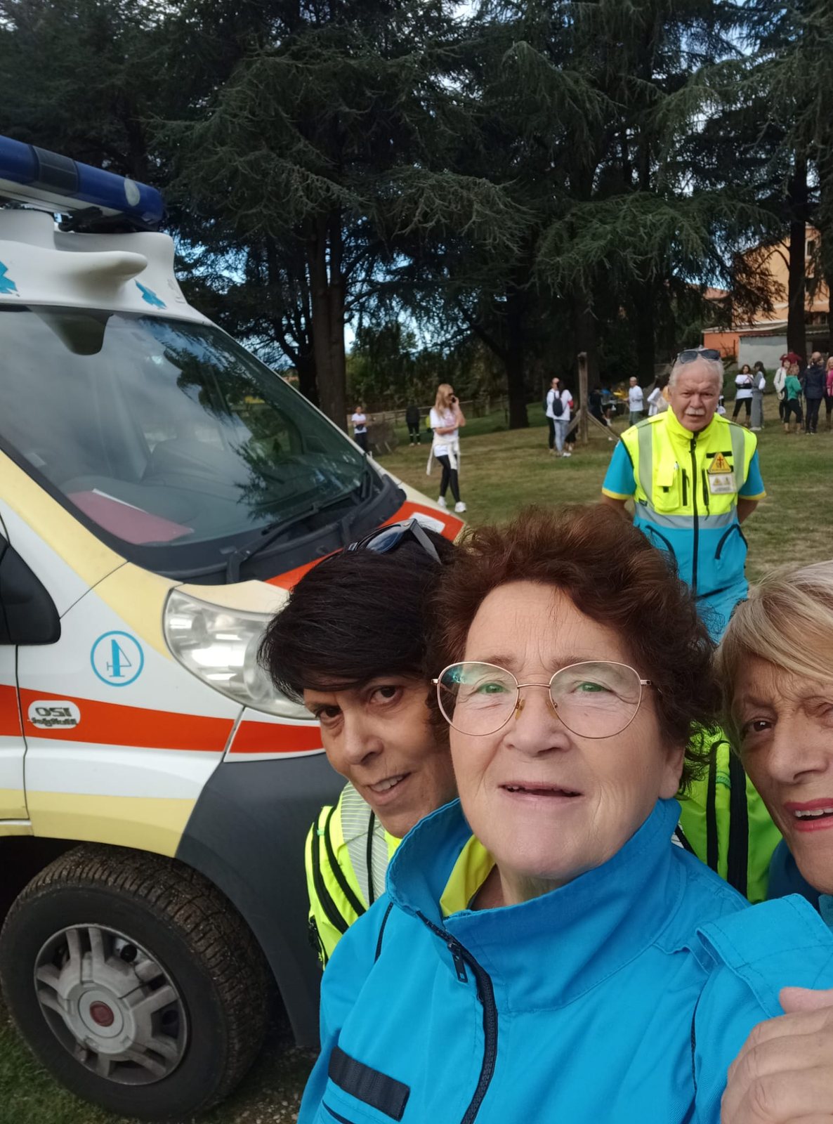 Selfie di tre volontarie della Misericordia accanto all'ambulanza ai Campi di Annibale, con un volontario in divisa e i partecipanti sul prato alle spalle