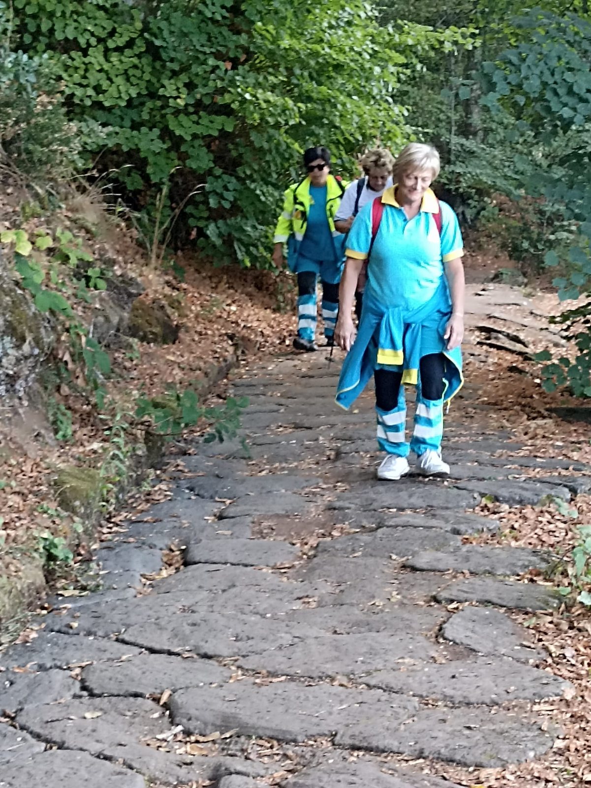 Volontarie della Misericordia in salita sulle pietre della Via Sacra, tra le foglie cadute e il verde del bosco