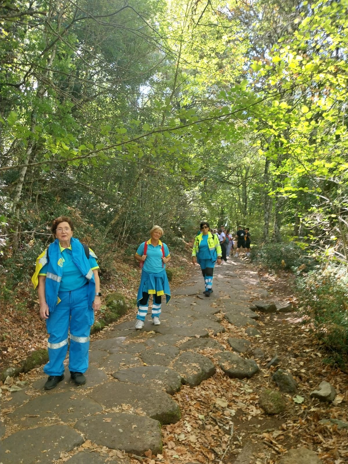 Tre volontarie della Misericordia in divisa azzurra camminano sull'antico basolato romano della Via Sacra, nel bosco di Monte Cavo, seguite dagli altri partecipanti