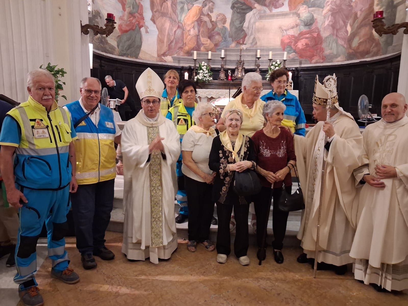 Foto di gruppo davanti all’altare della chiesa di Santa Maria Assunta in Cielo ad Ariccia: i volontari della Misericordia in divisa gialla e azzurra, i celebranti in paramenti bianchi con la mitria e alcune parrocchiane con il foulard giallo