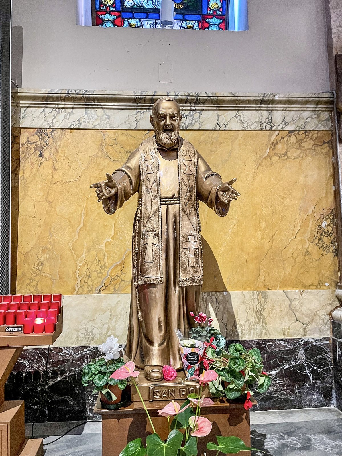 Statua in bronzo di San Pio da Pietrelcina con le braccia aperte e la stola sacerdotale, davanti a una parete di marmo giallo, con fiori e lumini ai piedi