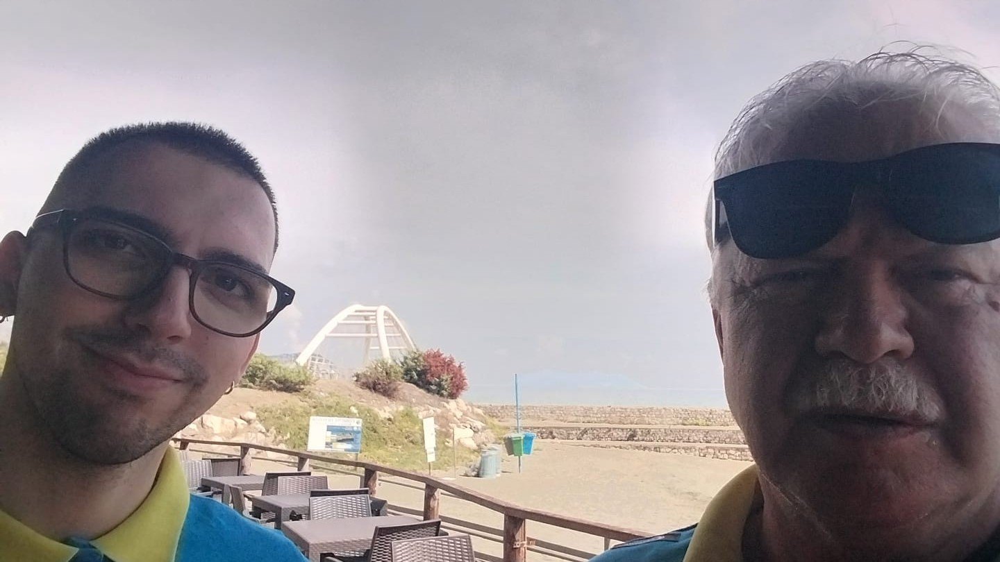 I due volontari della Misericordia di Ariccia in polo gialla e azzurra si fanno un selfie sul lungomare, con il mare e la spiaggia alle spalle, al termine del trasporto sanitario a Fondi