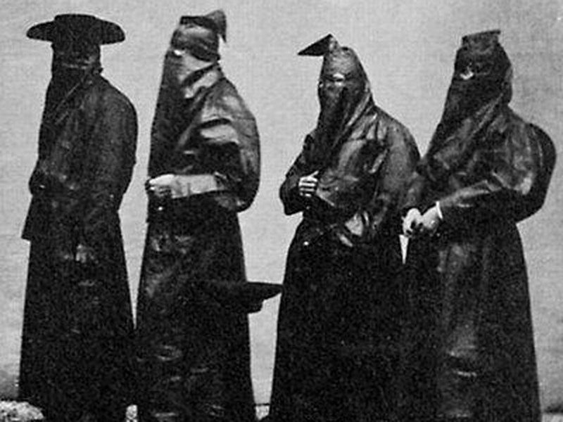 Fotografia in bianco e nero: quattro confratelli in cappa e buffa; il primo porta il cappello nero a falde larghe sopra la buffa, gli altri il sanrocchino lucido sulle spalle