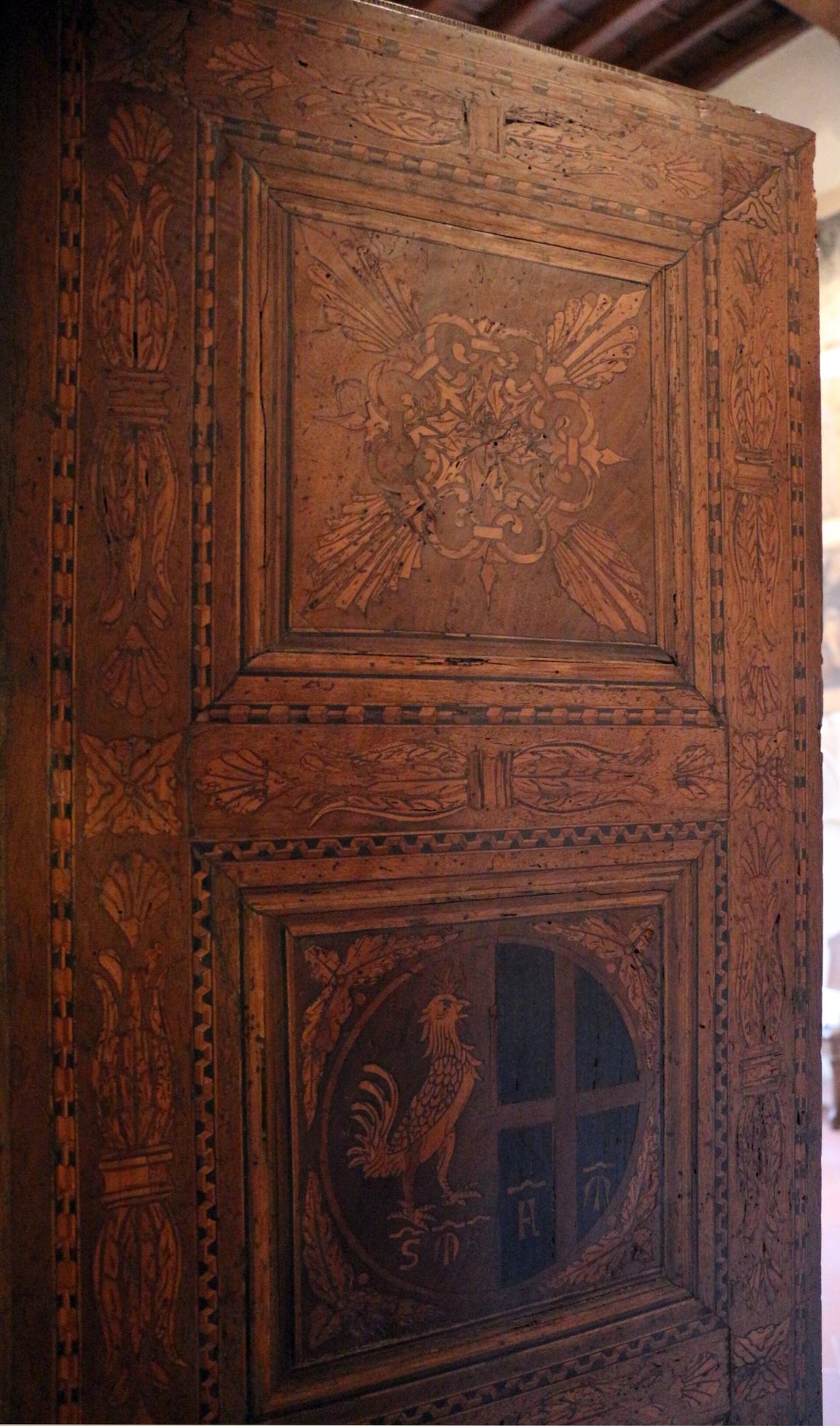 Porta in legno intarsiato; nel riquadro in basso, dentro un tondo, il gallo del Bigallo accanto alla croce con le lettere F e M